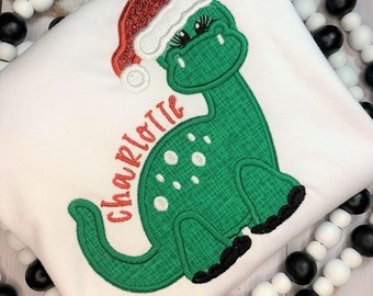 May include: White long-sleeved shirt with a green dinosaur wearing a red Santa hat. The dinosaur has white spots and the name "Charlotte" is embroidered on the shirt.