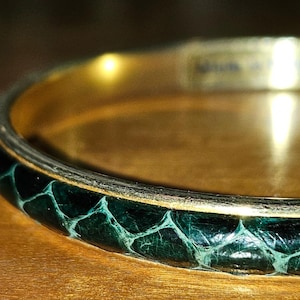 May include: Close-up of a gold bangle bracelet with a dark green snakeskin pattern. The bracelet has a gold interior and exterior trim. The snakeskin pattern is a dark teal color with a lighter teal pattern.