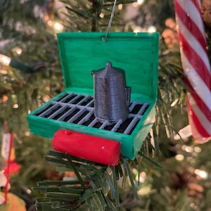 Pode incluir: Um enfeite de Natal impresso em 3D verde e vermelho em forma de cafeteira de acampamento. O enfeite tem uma tampa que se abre para revelar uma cafeteira prateada no interior.