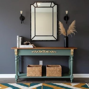 May include: A teal and brown console table with two woven baskets, a decorative mirror, and a black vase with dried plants. The table has gold accents and is against a dark gray wall.
