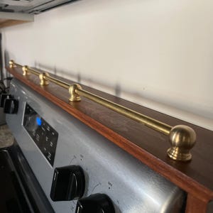 May include: A custom-made wooden shelf with a brass rail, designed to fit above a stove. The shelf has a dark wood finish with a brass rail and decorative brass ball finials. The stove is stainless steel with black knobs.