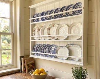 May include: A white plate rack filled with decorative plates. The plates have blue and white patterns, floral designs, and plain white surfaces. The rack is mounted on a wall above a wooden countertop with a bowl of lemons and a potted plant.