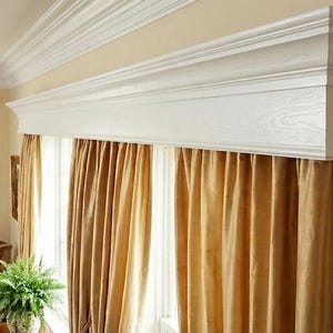 May include: A room interior featuring a white crown molding and a matching window valance. Gold-colored curtains hang below the valance. A fern plant sits in front of the window, and a brown armchair is visible.