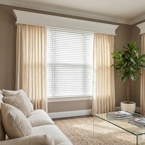 May include: A living room scene featuring a window with white blinds, framed by sheer beige curtains. A sofa with beige cushions and decorative pillows is in the foreground. A potted plant and glass coffee table complete the room's decor.