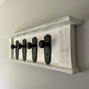 May include: White wooden shelf with a distressed finish, featuring five black antique door knobs as hooks. The shelf is mounted on a white wall.