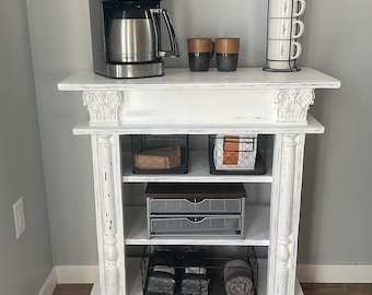 May include: White faux fireplace coffee station with a coffee maker, mugs, and storage baskets. The coffee station has three shelves and is decorated with a distressed white paint finish.