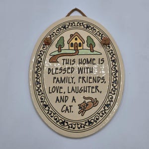 May include: Oval ceramic wall plaque with a cream background and a decorative border. The plaque features a house, trees, and a cat illustration. Text reads: "THIS HOME IS BLESSED WITH FAMILY, FRIENDS, LOVE, LAUGHTER, AND A CAT."