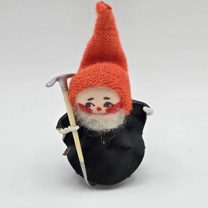 May include: A vintage gnome figurine with a red knit cap and a black body. The gnome has a white beard, rosy cheeks, and holds a silver and wooden pickaxe. The figurine is a charming piece of holiday decor.