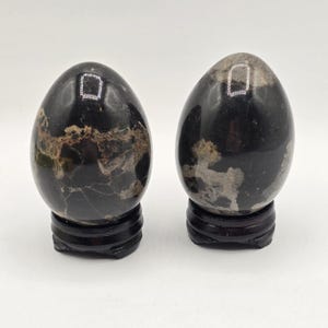May include: Two polished, egg-shaped stones with a smooth, glossy black surface featuring irregular, light brown and grey veining. Each stone is securely mounted on a circular, dark brown wooden display stand with a tiered base design.