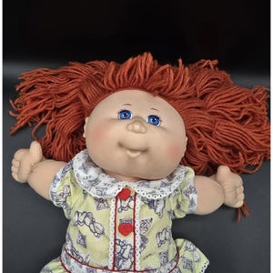 May include: A vintage doll with a bald head, blue eyes, and a smiling expression. The doll has red yarn hair and is wearing a yellow dress with a white collar and red heart-shaped buttons. The doll is lying down with its arms raised.