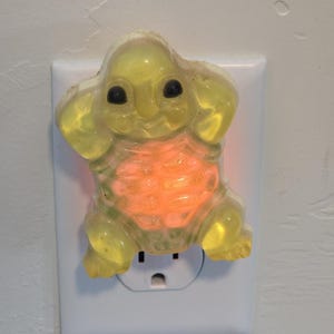 May include: A yellow night light shaped like a turtle, plugged into a white wall outlet. The turtle has black eyes and a glowing orange center. The night light is made of a translucent material.