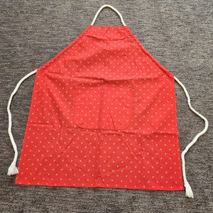 May include: Red apron with white polka dots and a front pocket. The apron has a white rope neck strap and waist ties. The apron is made of fabric and is designed for use in the kitchen or for crafting.