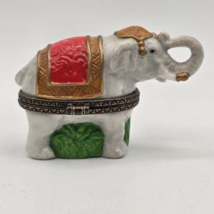 May include: A white ceramic elephant figurine with a red and gold saddle. The elephant has a raised trunk and a decorative gold headpiece. The base of the elephant is green, and the figurine is designed to open.