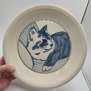May include: A cream-colored ceramic bowl with a hand-painted illustration of a cat in shades of blue. The cat is lying down, and the bowl has a simple, rounded design. The cat illustration is centered in the bowl.