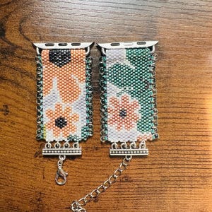 May include: Two beaded watch straps featuring floral patterns in peach, white, green, and black. Each strap has silver-tone metal connectors at the top and a silver-tone metal clasp with a chain extender at the bottom, finished with clear beads.