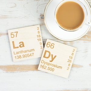May include: Two wooden coasters featuring the chemical element symbols and names for Lanthanum (La) and Dysprosium (Dy). The coasters are displayed on a white wooden surface with a white saucer and a cup of coffee.