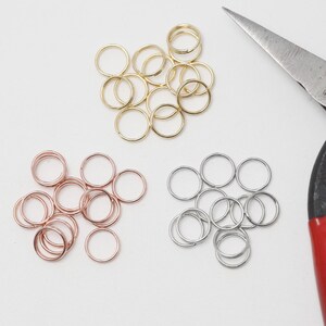 Op de afbeelding: Een assortiment gouden, roségouden en zilveren sprongringen ligt op een witte ondergrond. Een rood-handvat metalen schaar is in de buurt geplaatst, wat op knutselbenodigdheden duidt.