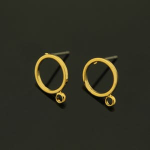 Peut inclure: Une paire de boucles d'oreilles dorées. Chaque boucle d'oreille présente un design circulaire avec une petite boucle en bas. Les boucles d'oreilles sont posées sur un fond sombre, soulignant leur éclat métallique.