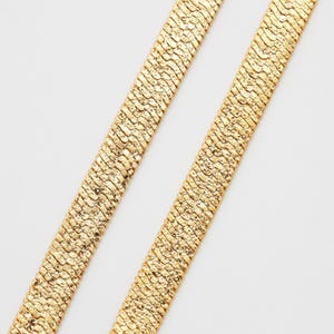 May include: Two gold-colored, textured, flat chain necklaces are displayed against a white background. The chains have a woven pattern, and the gold color has a slightly aged appearance. The chains are positioned diagonally.