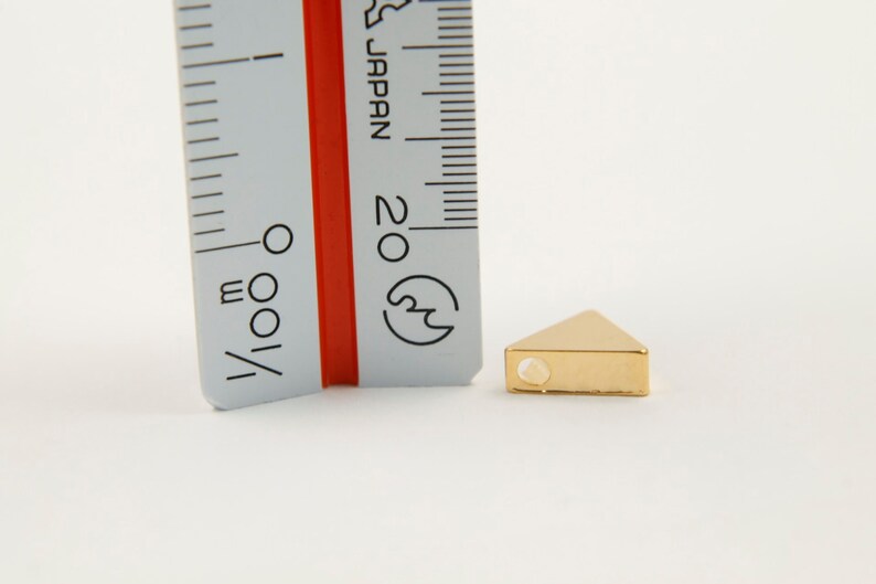 May include: A gold triangle-shaped bead with a hole in the centre. The bead is approximately 1 cm wide and is shown next to a ruler.