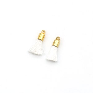 May include: Two white tassel charms with gold metal caps. The tassels are made of soft, white yarn and have a delicate, fringed appearance.