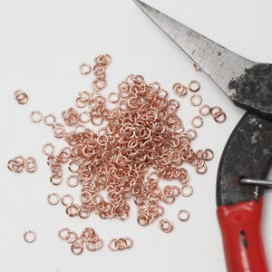 Op de afbeelding: Een verzameling kleine, koperkleurige sprongringen verspreid op een wit oppervlak, naast een paar gebruikte metalen tangen met rode handvatten. De tang is gedeeltelijk zichtbaar, wat wijst op een knutsel- of sieradenmaakthema.
