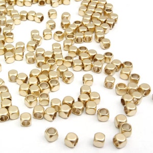 Puede incluir: Una gran cantidad de cuentas de metal de color dorado en forma cuadrada. Las cuentas están esparcidas sobre una superficie blanca.