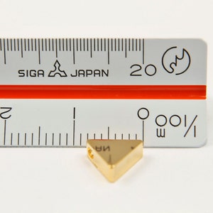 May include: A gold-coloured triangle-shaped pendant with a hole in the top. The pendant is measured against a ruler, showing it is approximately 3.8 cm in length.