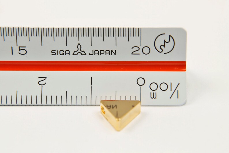 May include: A gold-coloured triangle-shaped pendant with a hole in the top. The pendant is measured against a ruler, showing it is approximately 3.8 cm in length.