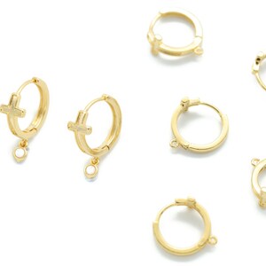 Peut inclure: Six boucles d'oreilles créoles dorées avec un pendentif en forme de croix et une petite pierre transparente. Les boucles d'oreilles sont sur un fond blanc.