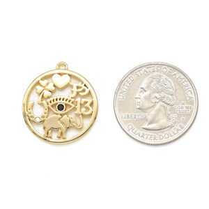 May include: Gold-colored charm with a circular design featuring a heart, four-leaf clover, horseshoe, elephant, and the number 13. A U.S. quarter is next to the charm for size comparison. The charm has a small loop at the top for hanging.