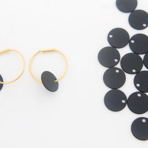 Op de afbeelding: Een paar gouden creolen met zwarte ronde bedels. De oorbellen liggen op een witte achtergrond. Er liggen verschillende zwarte ronde bedels verspreid op de rechterkant van de afbeelding.