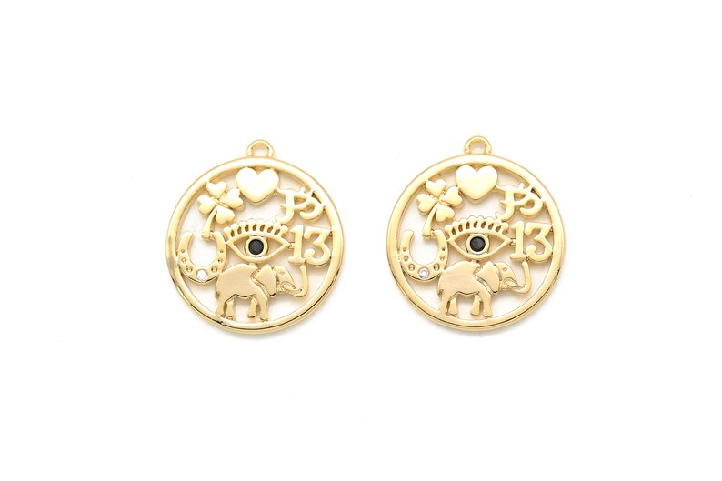 May include: Two gold-colored charm pendants with a circular design. Each pendant features a heart, a four-leaf clover, an eye, a horseshoe, an elephant, and the number 13. The eye has a black center. The pendants are on a white background.