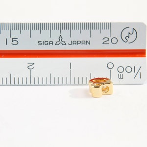 Puede incluir: Un charm de metal dorado en forma de la letra "Q". El charm mide aproximadamente 1,2 cm de alto y est&aacute; sobre una superficie blanca junto a una regla.