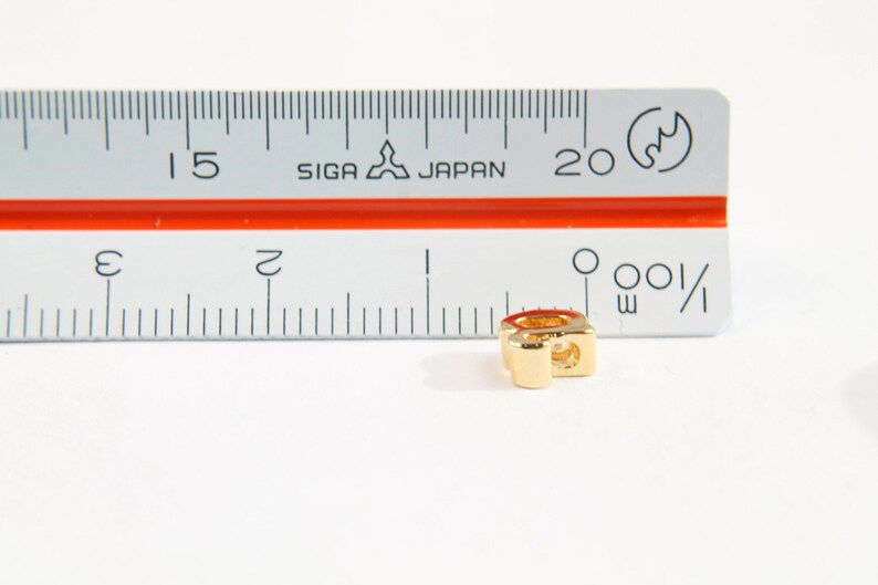 Puede incluir: Un charm de metal dorado en forma de la letra "Q". El charm mide aproximadamente 1,2 cm de alto y est&aacute; sobre una superficie blanca junto a una regla.