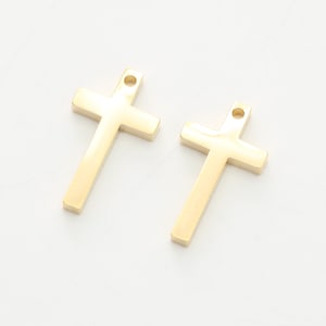 Puede incluir: Dos dijes de cruz de metal de color dorado con un pequeño agujero en la parte superior para sujetar a un collar o pulsera.