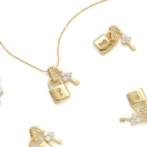Puede incluir: Collar dorado con un colgante de candado y un dije de llave, ambos adornados con pequeños cristales brillantes. El collar y los dijes se colocan sobre un fondo blanco, creando una estética brillante y elegante.