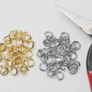 Op de afbeelding: Een bovenaanzicht toont twee stapels metalen verbindingsringen, één goudkleurig en de andere zilverkleurig. Een roestige tang met rode handvatten op de achtergrond, wat duidt op sieraden maken of knutselbenodigdheden.