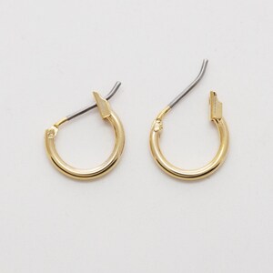 May include: A pair of small, gold-coloured hoop earrings. Each earring has a curved, circular design with a post and clasp closure. The earrings are displayed on a white surface.