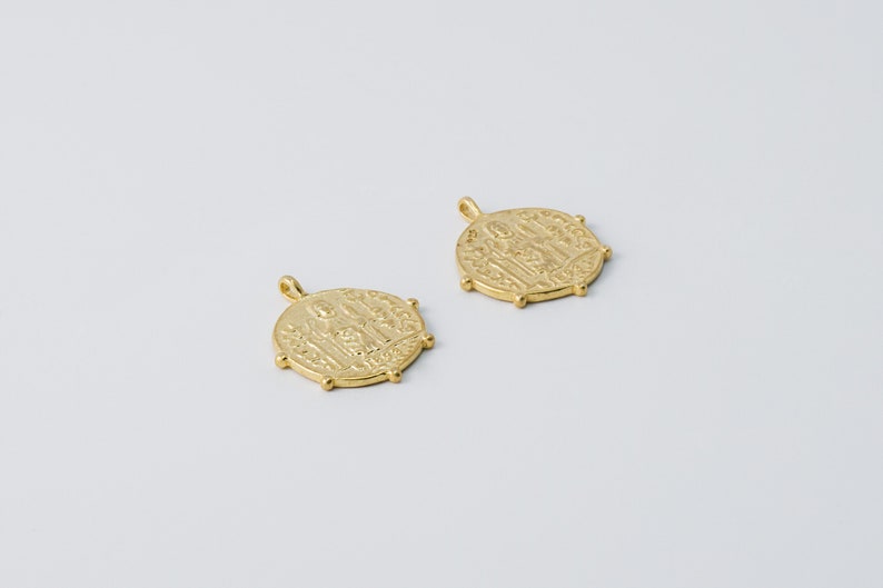 May include: Two gold-toned metal pendants with a raised design of figures. The pendants are round and have a loop at the top for attaching to a chain.