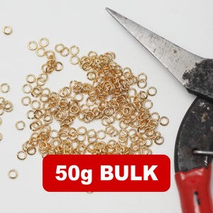 Op de afbeelding: Een verzameling kleine, goudkleurige verbindingsringen verspreid op een witte ondergrond, met een tang op de achtergrond. Een rode rechthoek met de tekst "50g BULK" is zichtbaar. Ideaal voor het maken van sieraden en knutselen.
