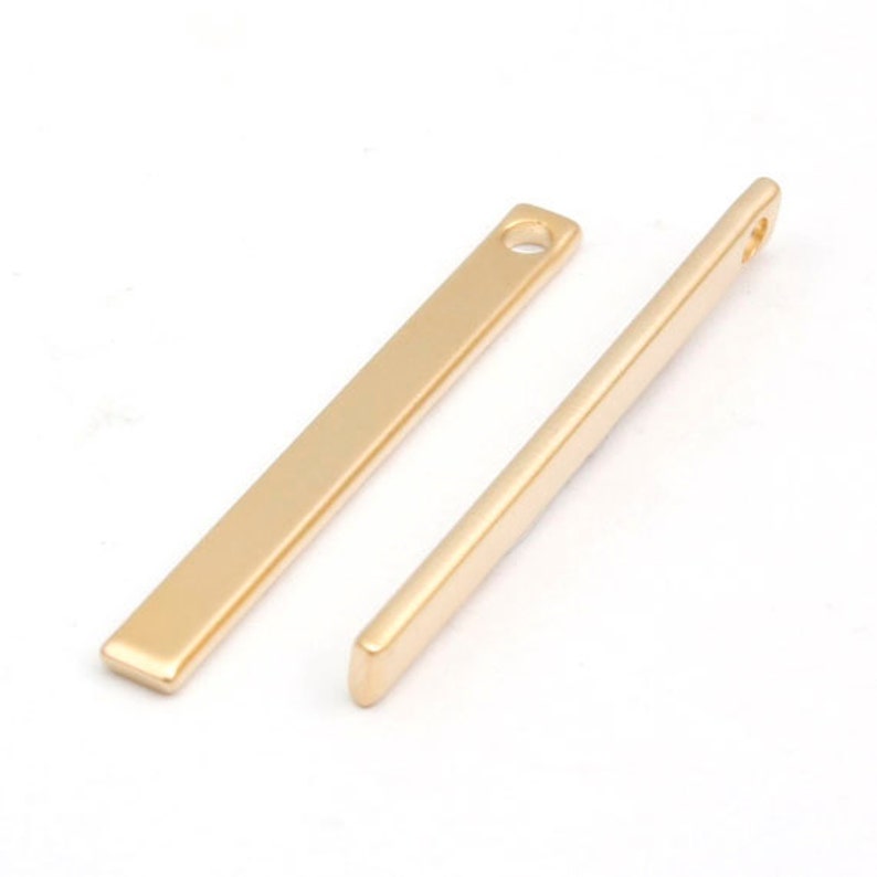 May include: Two gold-colored metal bar pendants with a small hole at the top. The bars are smooth and have a rectangular shape.