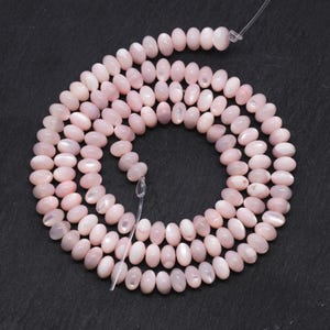 Peut inclure: Un fil de perles ovales rose clair, enfilées ensemble avec une fine ficelle transparente. Les perles sont disposées en spirale sur une surface gris foncé. Les perles ont un lustre subtil.
