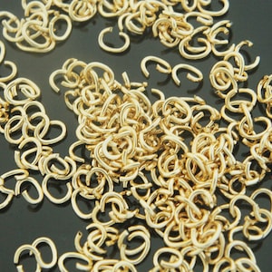 May include: A collection of small, gold-colored open jump rings scattered across a dark surface. The rings are in a variety of shapes and sizes, with a shiny, metallic finish. These jewelry findings are ideal for crafting and jewelry making.