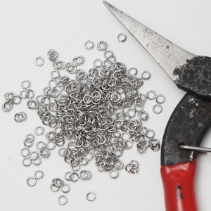 Op de afbeelding: Een close-up toont een stapel kleine, zilveren metalen sprongringen naast een tang met rode handvatten. De tang is gedeeltelijk zichtbaar, met de bladen en een deel van het handvat in beeld. De sprongringen zijn voor het maken van sieraden.