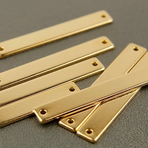 Könnte beinhalten: Eine Gruppe von sechs goldfarbenen Metallstangen mit abgerundeten Kanten und Löchern an jedem Ende. Die Stangen sind in einem zufälligen Muster auf einer grauen Oberfläche angeordnet.