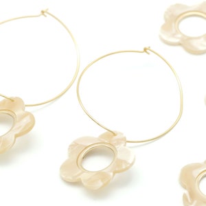Op de afbeelding: Een paar gouden creolen met een crèmekleurige bloemenhanger aan elke creool.