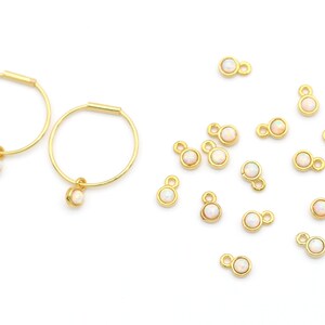 May include: Two gold hoop earrings with a small white opal charm dangling from each hoop. A collection of 14 gold charms with white opal stones are scattered around the earrings.