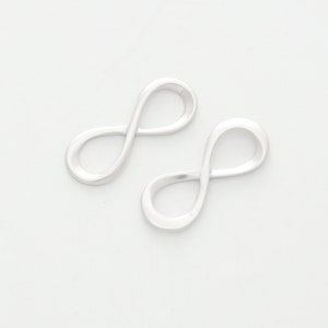 Może przedstawiać: Dwa srebrne charms w kształcie symbolu nieskończoności. Charms są gładkie i błyszczące.