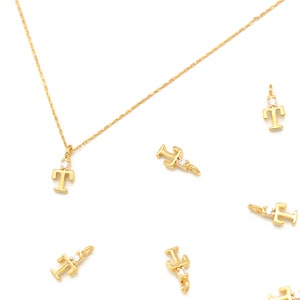 May include: A gold chain necklace with a gold letter T pendant with a small clear stone. The image also shows several other gold letter T pendants with clear stones.